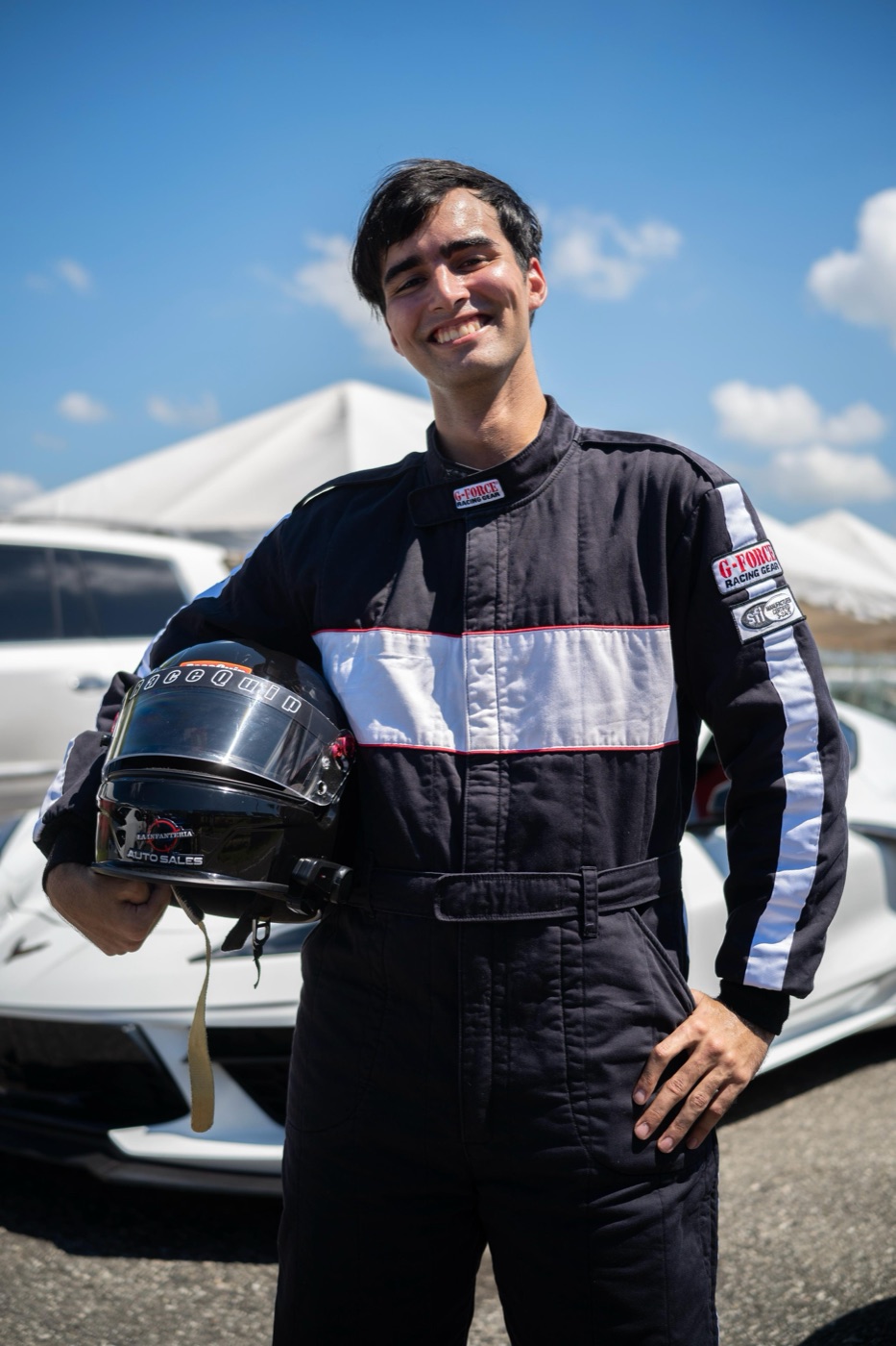 Oliver Infante con traje de competencia, sosteniendo el casco frente a un Corvette en el circuito.