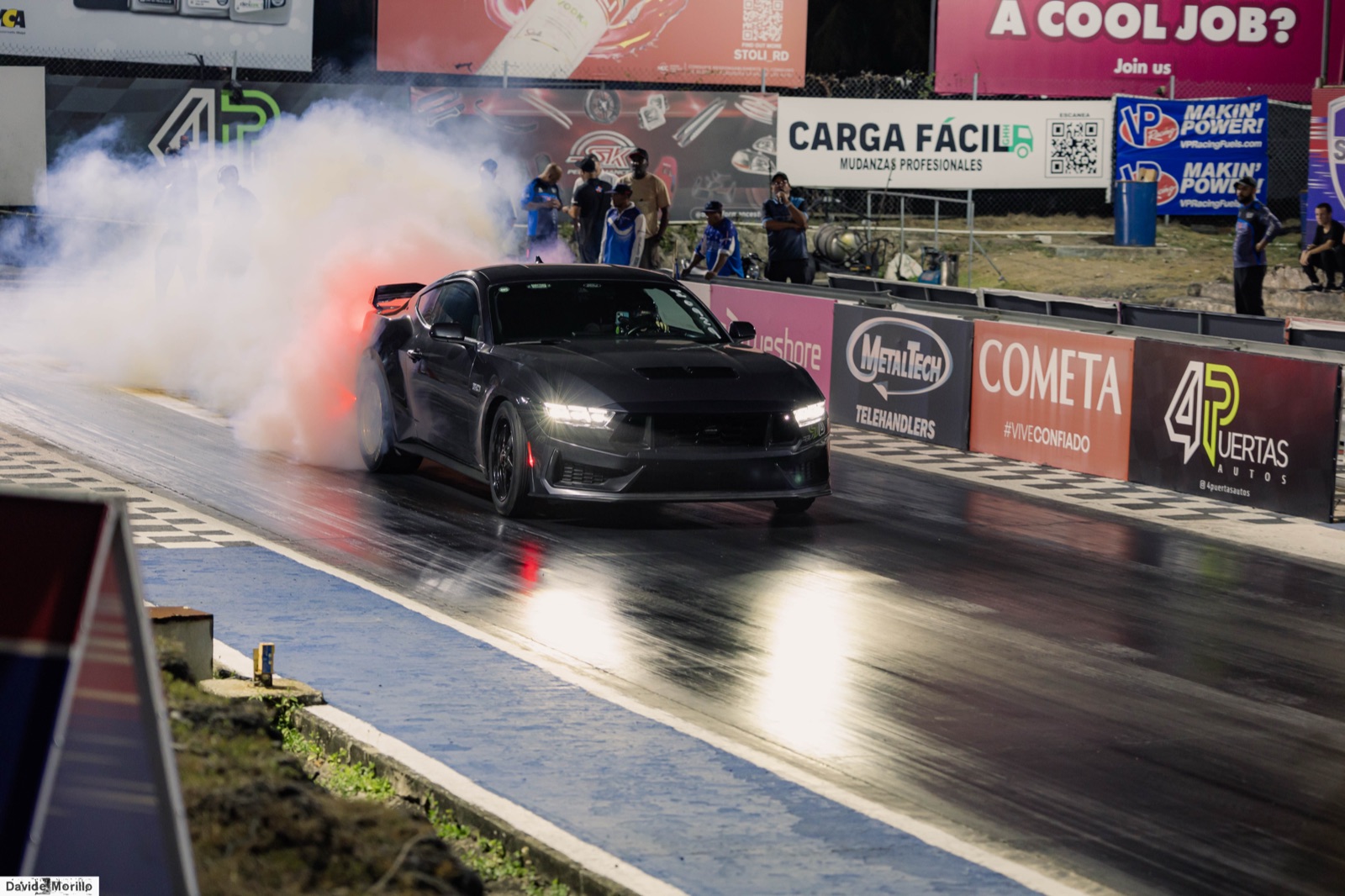 Ford Mustang negro haciendo el burnout previo a una pasada de cuarto de milla, con la pista mojada y humo de neumáticos.